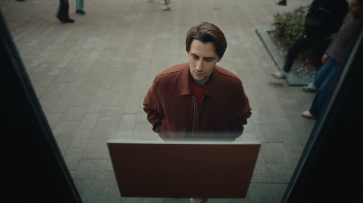 Young white man in a red jacket standing in front of a window, looking at a sign hung in there