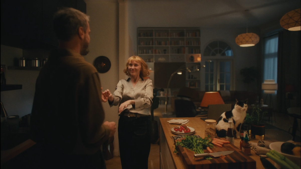 Young white woman with short red hair is dancing romantically with a man in a warmly lit kitchen. On the counter the food is prepared and a black-white cat is is sitting there too.